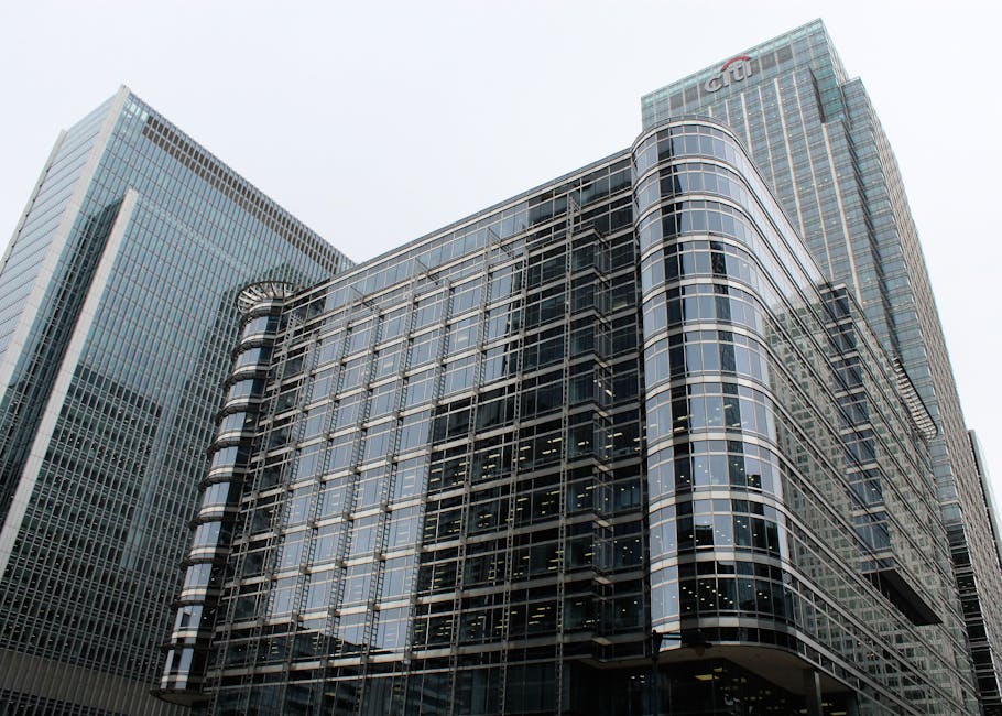 A modern multi-storey office building with a rounded corner, featuring extensive glass facades and metallic framing, located among other high-rise structures in an urban financial district. The building's exterior includes large reflective windows that reveal interior office spaces, with some sections showing dark internal floors and ceiling fixtures. The surrounding environment consists of neighboring glass-covered skyscrapers, one with vertical lines of windows and another taller tower with a logo near the top. The sky above is overcast, providing diffuse lighting that slightly dulls reflections on the glass surfaces. The scene's clean and professional appearance aligns with the context of commercial property management and the potential for on-site waste or rubbish clearance, as London Rubbish Clearance offers services for such urban settings, facilitating alternative waste handling solutions away from municipal collection, if waste is visible nearby.