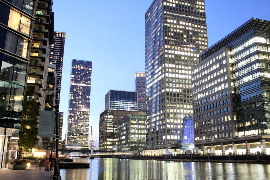 A daytime view of a modern urban waterfront with tall glass office buildings lining the promenade. The buildings feature reflective facades, with some showing illuminated windows and others reflecting the blue sky and nearby structures. In the foreground, part of a glass-fronted commercial building with a dark metal framework is visible on the left, and there are potted plants along the paved walkway. To the left, a street lamp and a vertical banner are installed along the pedestrian area. The waterway runs parallel to the buildings, with gentle ripples and a calm surface reflecting the ambient light. Overall, the scene depicts a contemporary financial district environment, which may relate to independent waste management or rubbish collection services for busy office buildings in the area, supported by the professional urban setting.